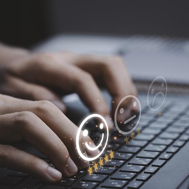 Hands typing on a keyboard. In front of the hands are a frown and a happy emoji.