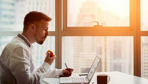 A young man eats an apple while working on his laptop.